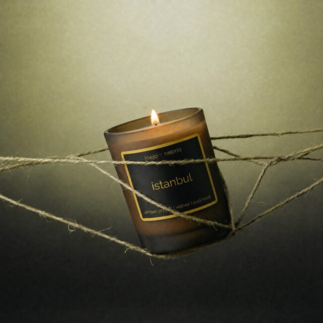 JoeJo + Nebria Istanbul luxury scented candle with amber crystal, vetiver, and oud musk fragrance notes, glowing within an artistic arrangement of intertwined natural rope against a warm neutral background.