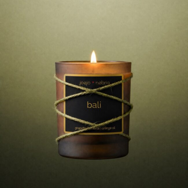 JoeJo + Nebria Bali luxury scented candle featuring grapefruit, neroli, and orange oil fragrance notes, wrapped in natural rope with a glowing flame against a soft green gradient background.