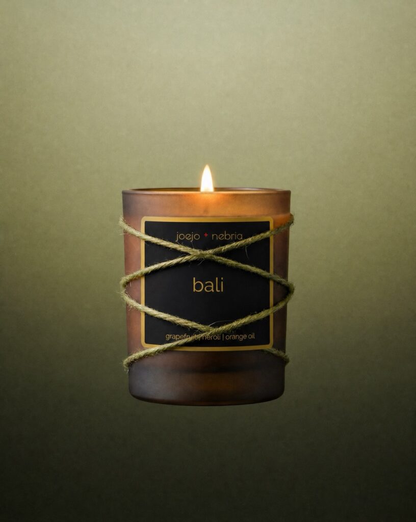 JoeJo + Nebria Bali luxury scented candle featuring grapefruit, neroli, and orange oil fragrance notes, wrapped in natural rope with a glowing flame against a soft green gradient background.