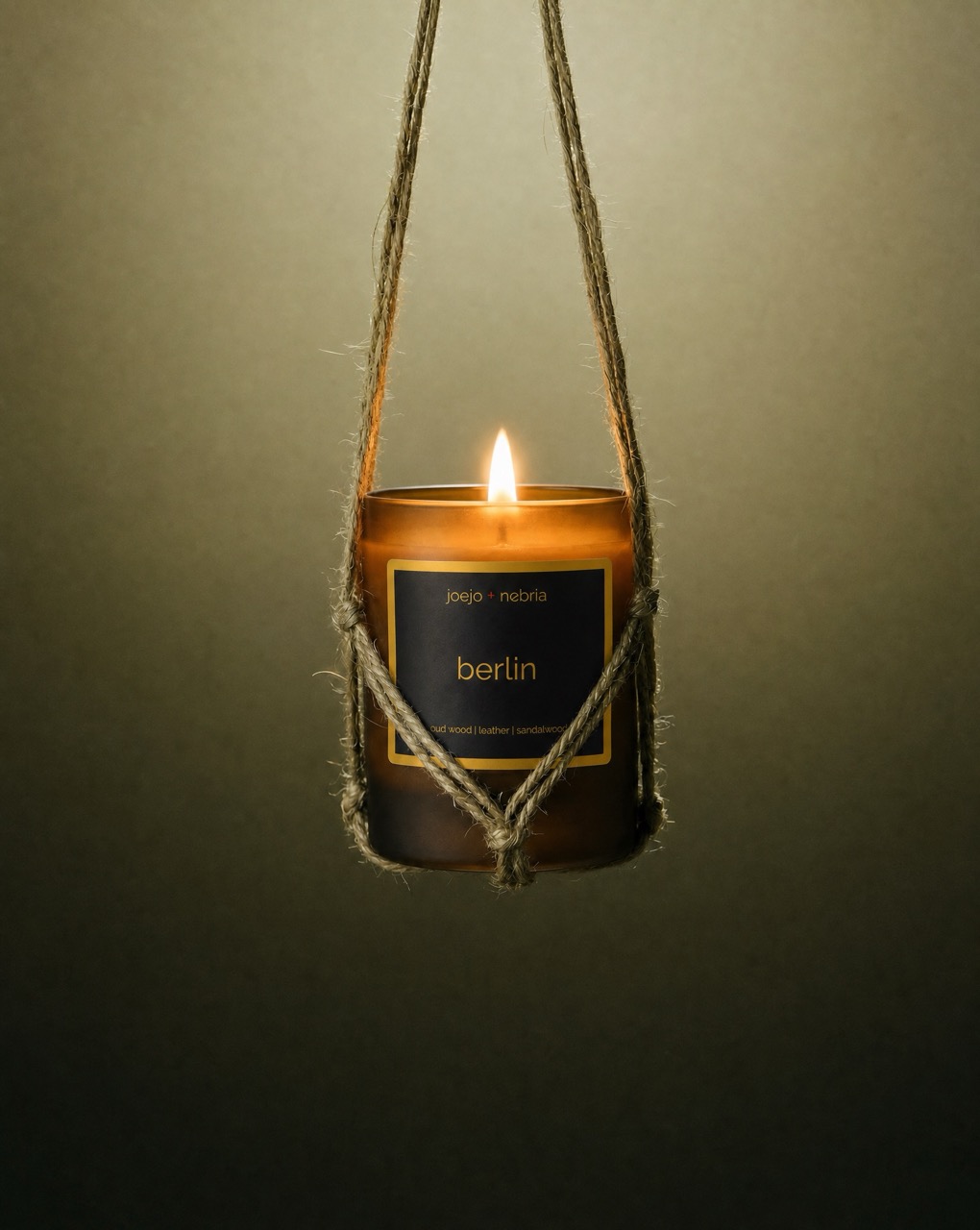 JoeJo + Nebria Berlin luxury scented candle with oud wood, leather, and sandalwood fragrance notes, suspended in a handcrafted rope holder with a glowing flame against a moody olive-toned background.