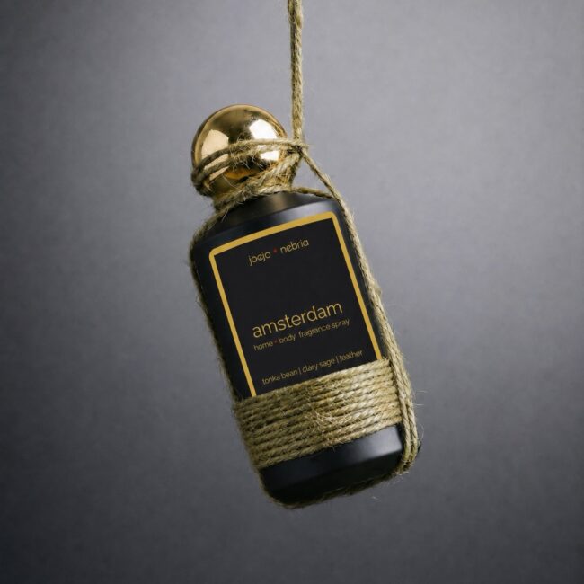 JoeJo + Nebria Amsterdam Home + Body Fragrance Spray featuring tonka bean, clary sage, and leather fragrance notes, displayed in a handcrafted rope holder with a gold cap against a dark textured background.