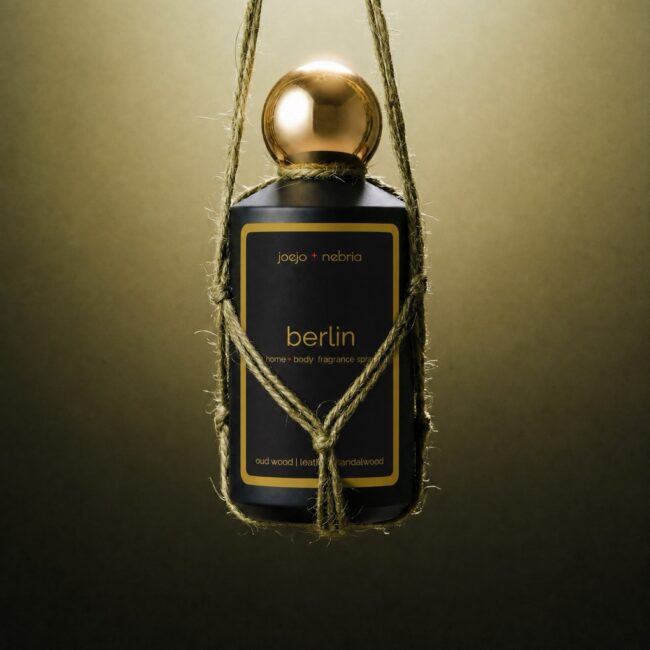 JoeJo + Nebria Berlin Home + Body Fragrance Spray featuring oud wood, leather, and sandalwood fragrance notes, suspended in a handcrafted rope holder with a gold cap against a moody olive-toned background.