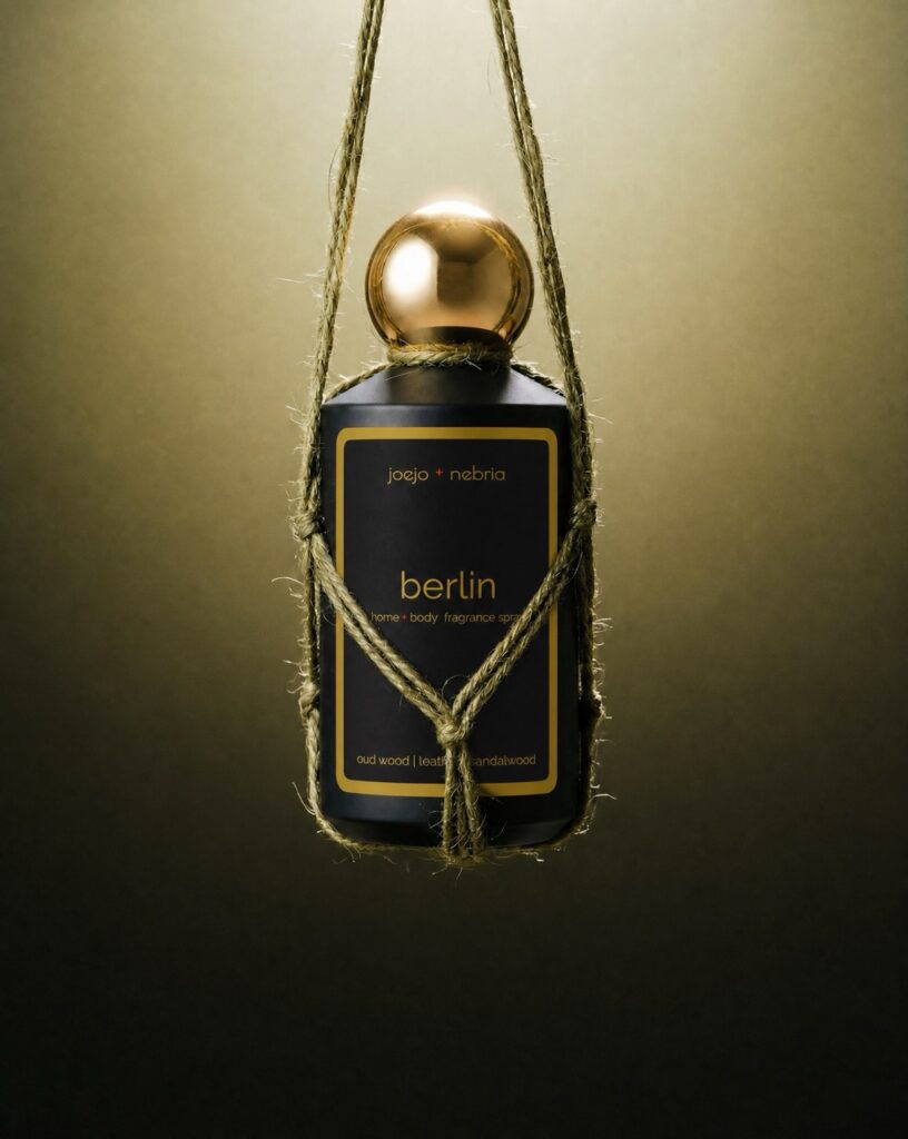 JoeJo + Nebria Berlin Home + Body Fragrance Spray featuring oud wood, leather, and sandalwood fragrance notes, suspended in a handcrafted rope holder with a gold cap against a moody olive-toned background.